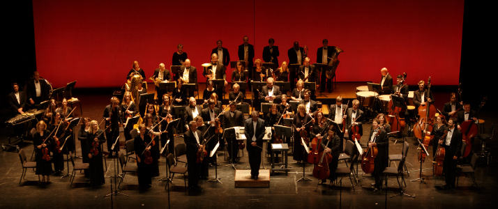 An entire orchestra posing for a picture in front of their chairs while holding their instruments