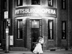 A black and white photo of a woman in a wedding dress and a man in a tux kissing in front of the Bitz Opera Factory