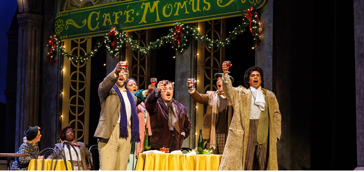 A woman in a blue robe sitting on a couch leans towards a man while looking heind herself with her head facing forward as a woman in a dress and apron stands in a doorway with her hand on the door. The man is wearing a barber's uniform and looks at the woman in blue