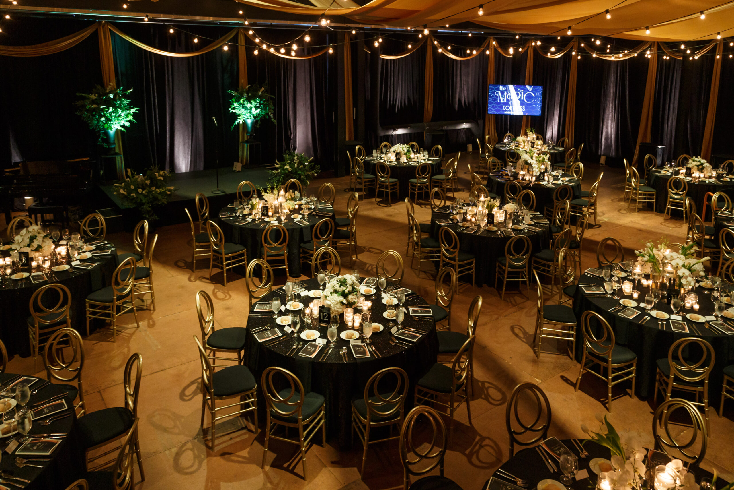 Round tables with black cloths, gold chairs, and white florals under string lights in the Bitz Opera Factory for Maecenas