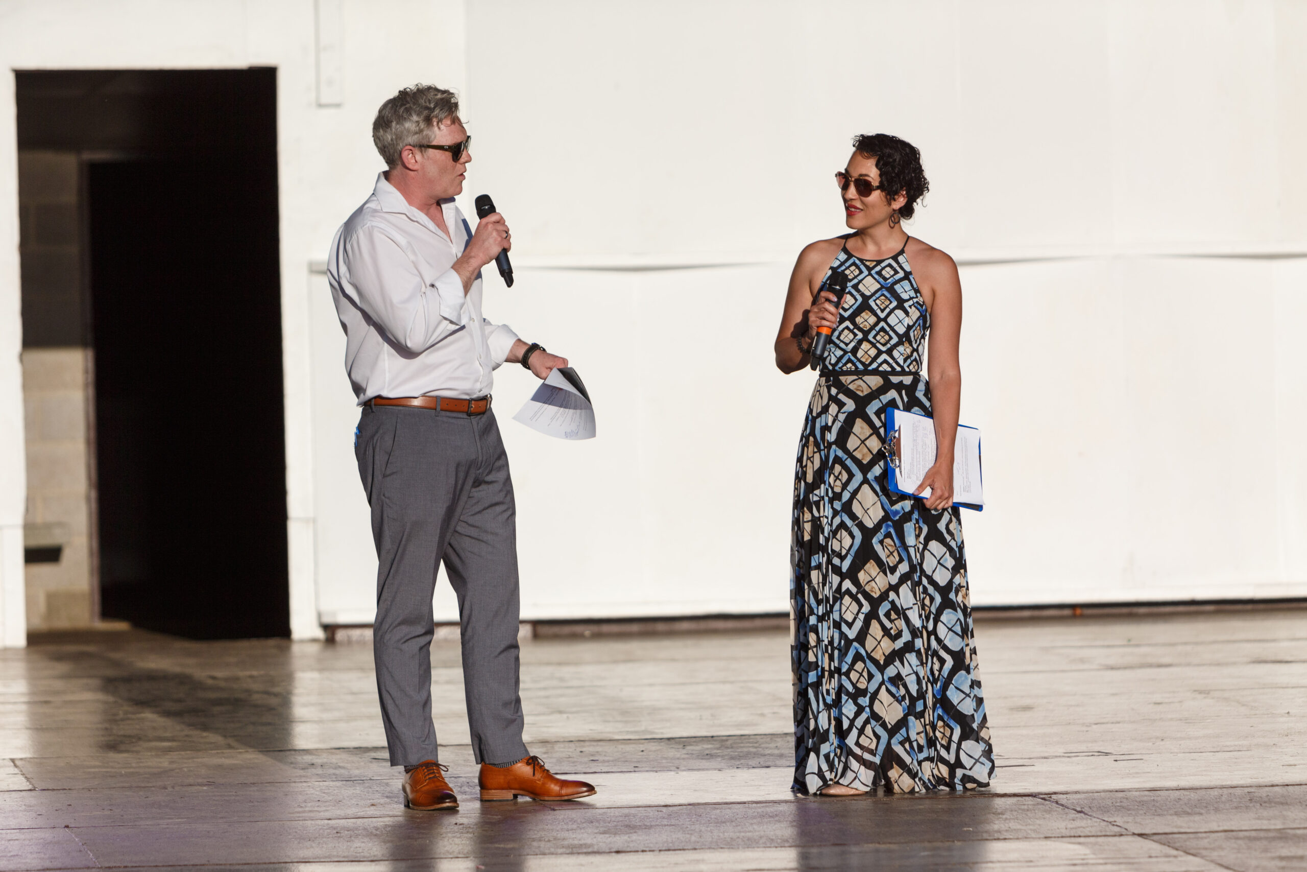 Two hosts with microphones, a man in white and a woman in a patterned gown, speak on the Hartwood Acres stage
