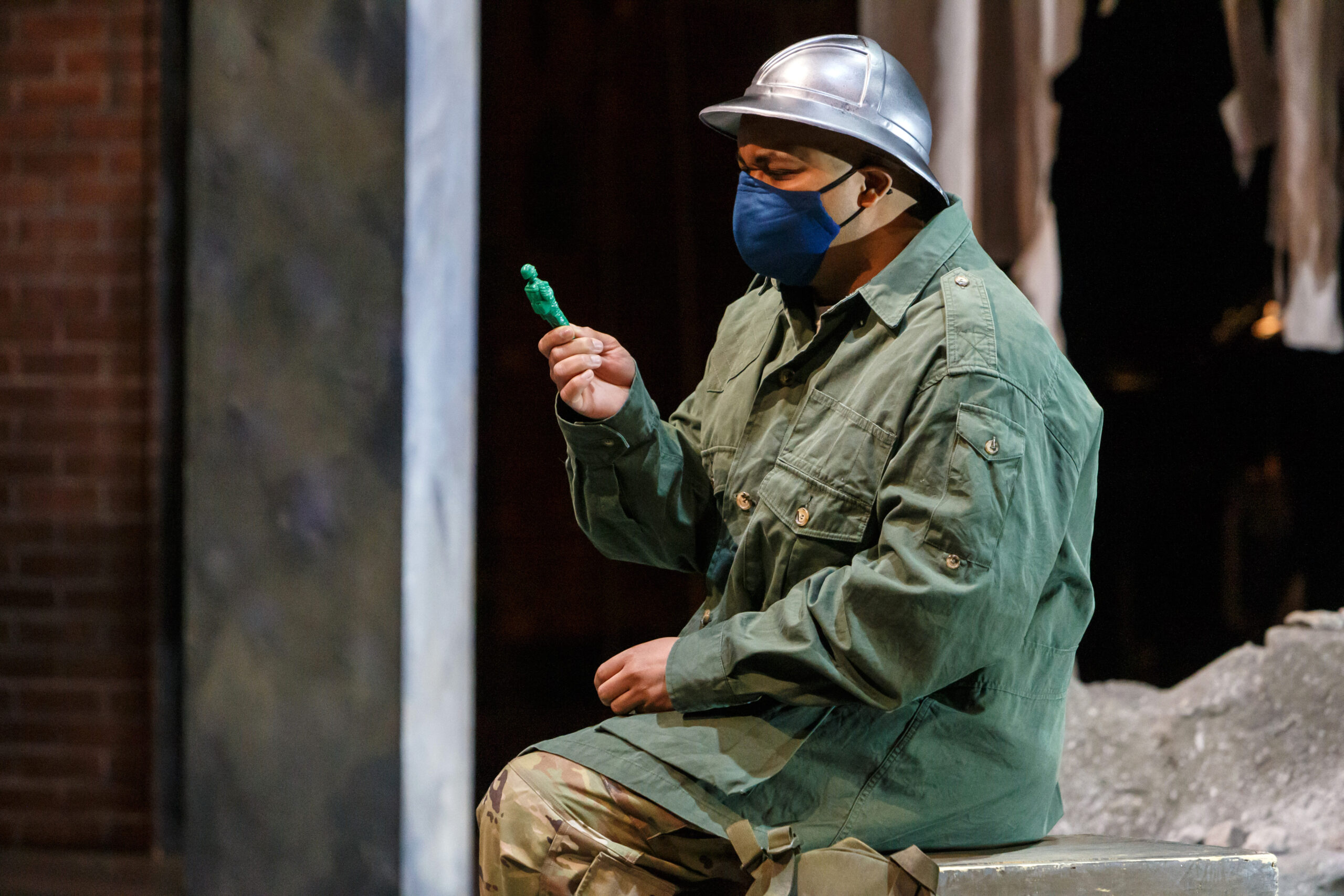 A masked soldier in a silver helmet and green fatigues studies a green toy soldier in Pittsburgh Opera's Soldier Songs, 2020