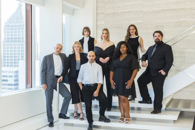 The 2024-25 Pittsburgh Opera Resident Artists pose on a staircase with the Downtown skyline in the windows behind