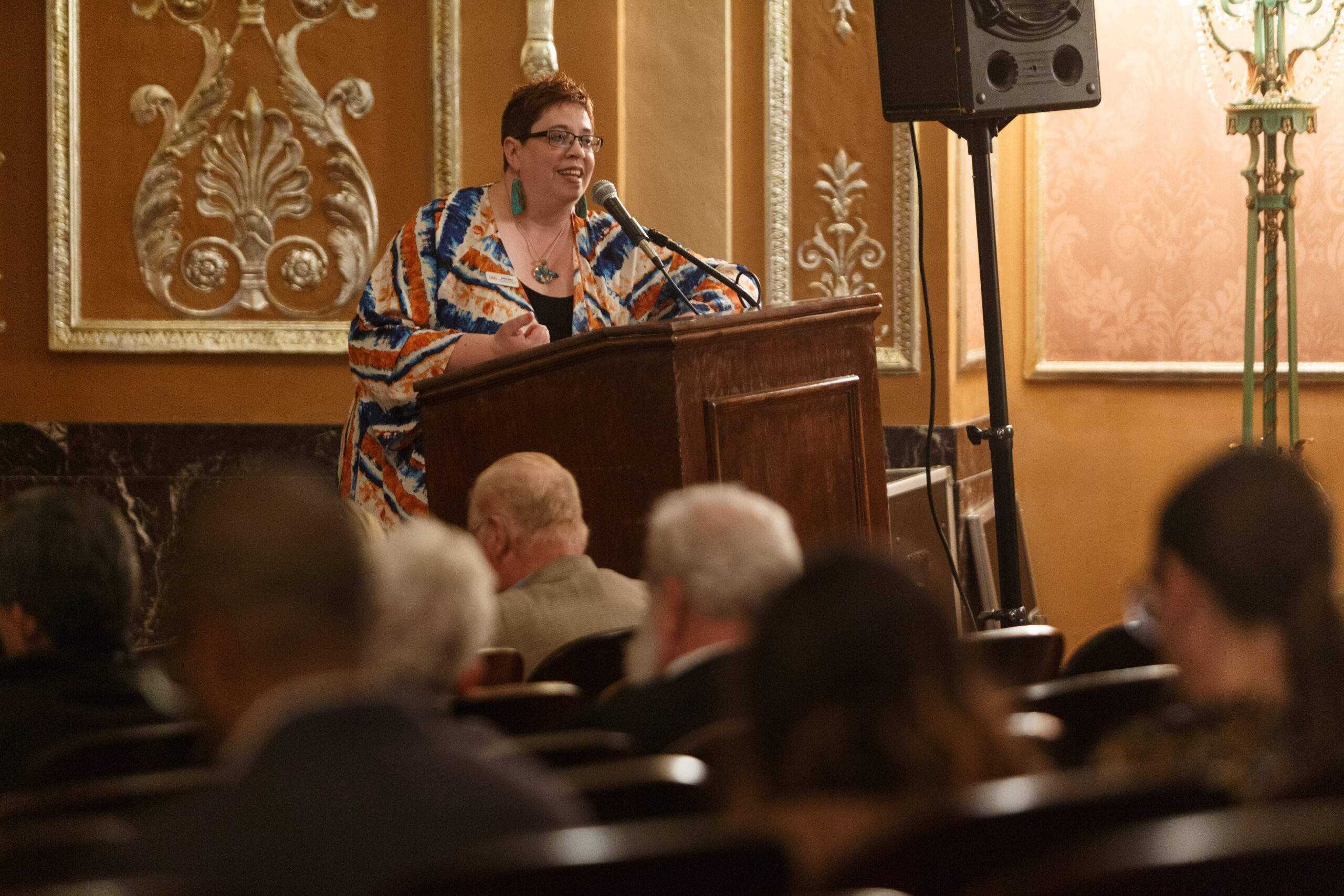 Kristin Gatch in a colorful patterned jacket speaks from a podium in the gilded Benedum Center before The Barber of Seville