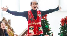 A singer in a holiday sweater vest spreads his arms before a Christmas tree at a Holiday Brown Bag concert