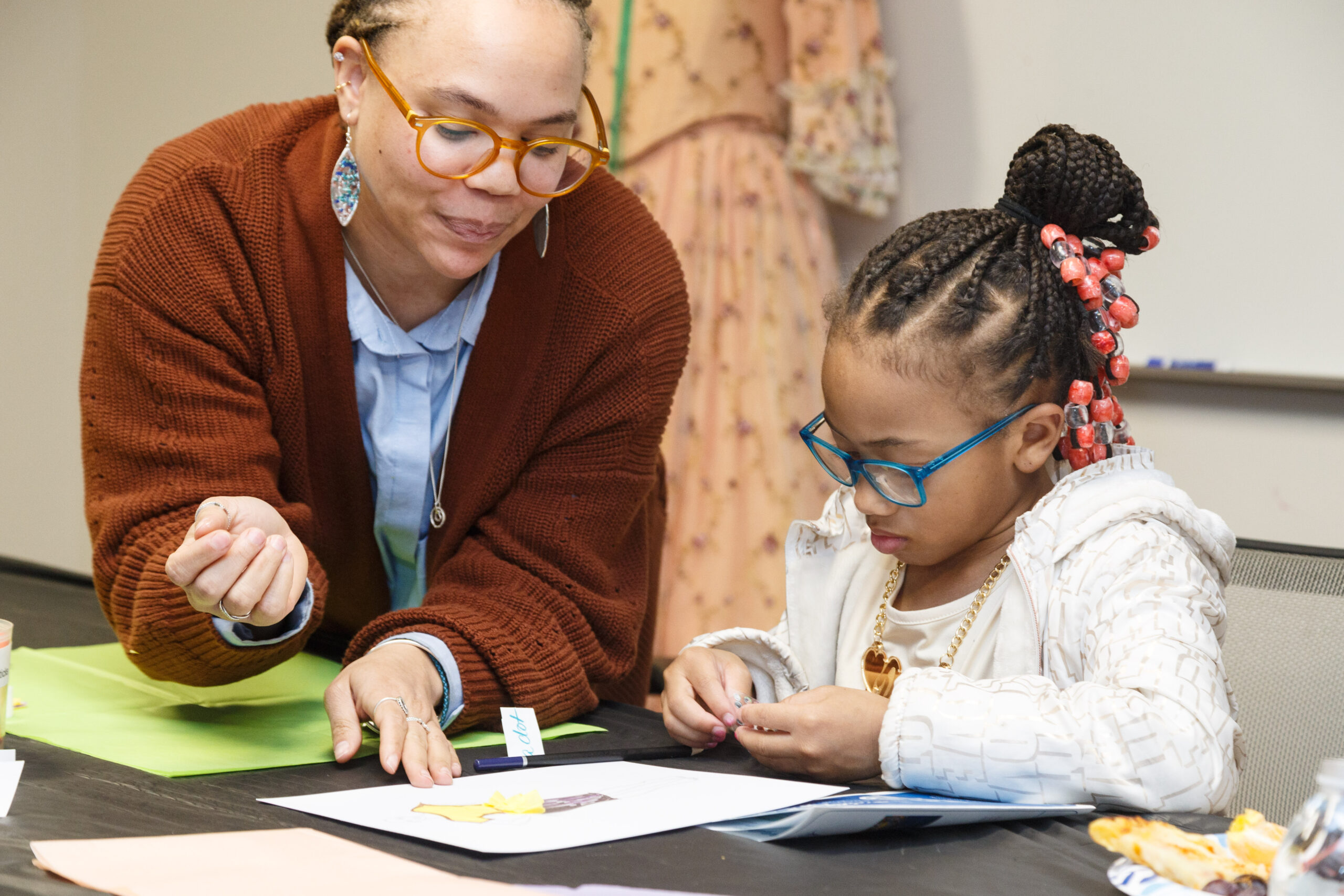 A woman in a rust cardigan helps a young girl in blue glasses with a craft at a Family Day table