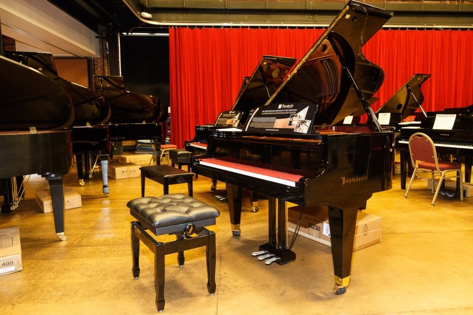 A Bosendorfer grand piano among rows of pianos before a red curtain at the Pittsburgh Opera piano sale