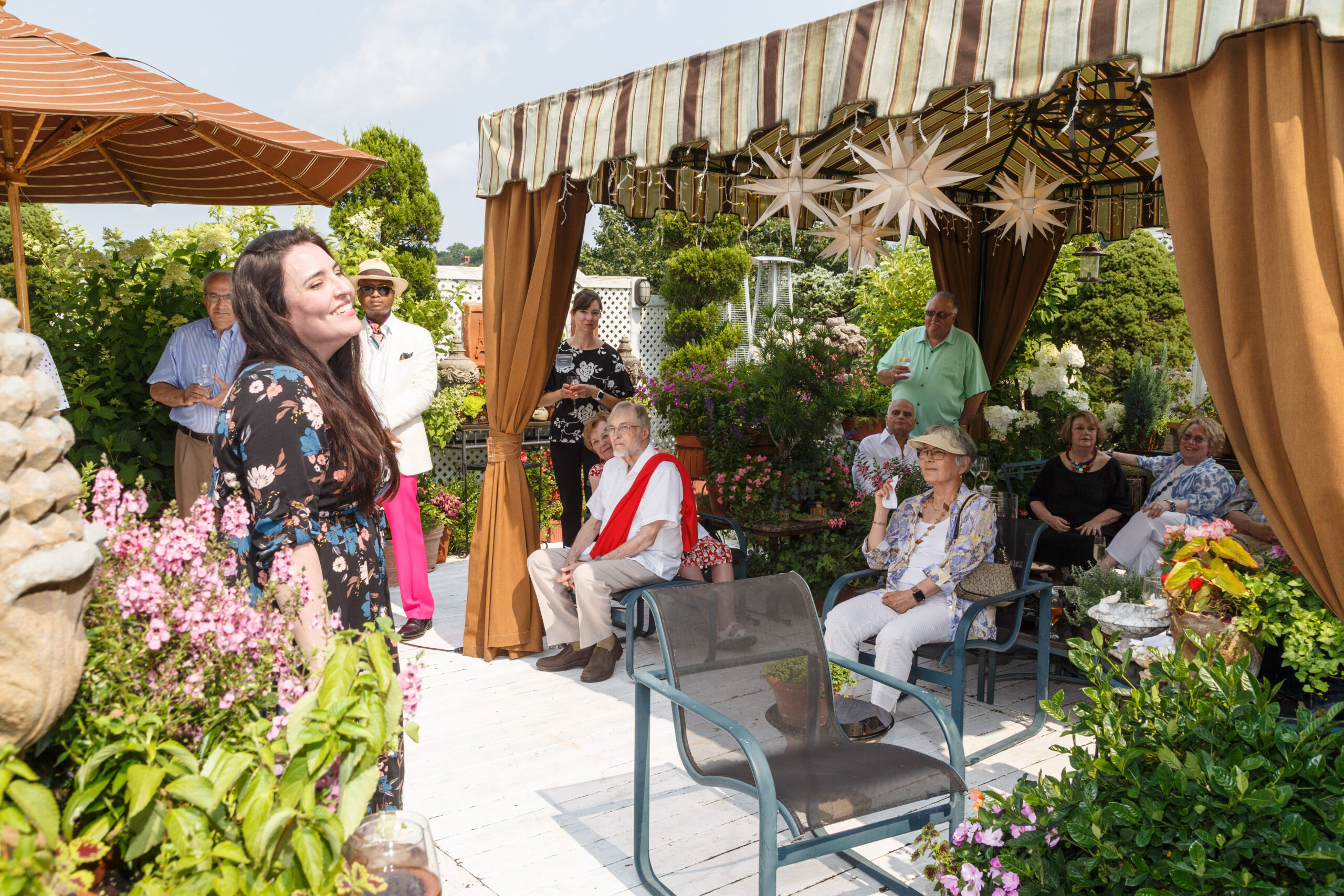 A young woman smiles before guests seated under a striped awning at a Friends of Pittsburgh Opera garden party