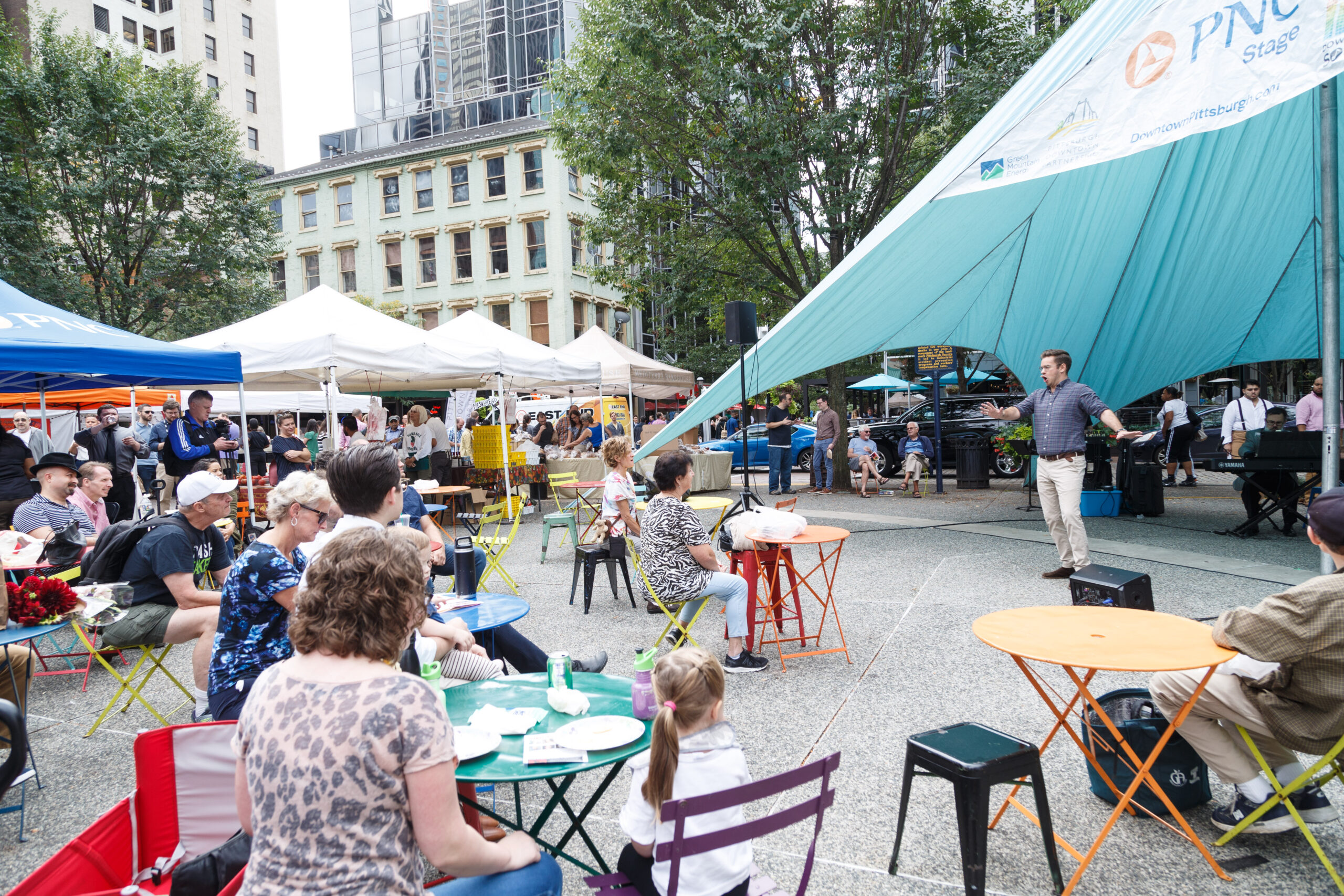 A Resident Artist sings under the teal PNC Stage canopy to a seated crowd at the Market Square farmers market, 2019