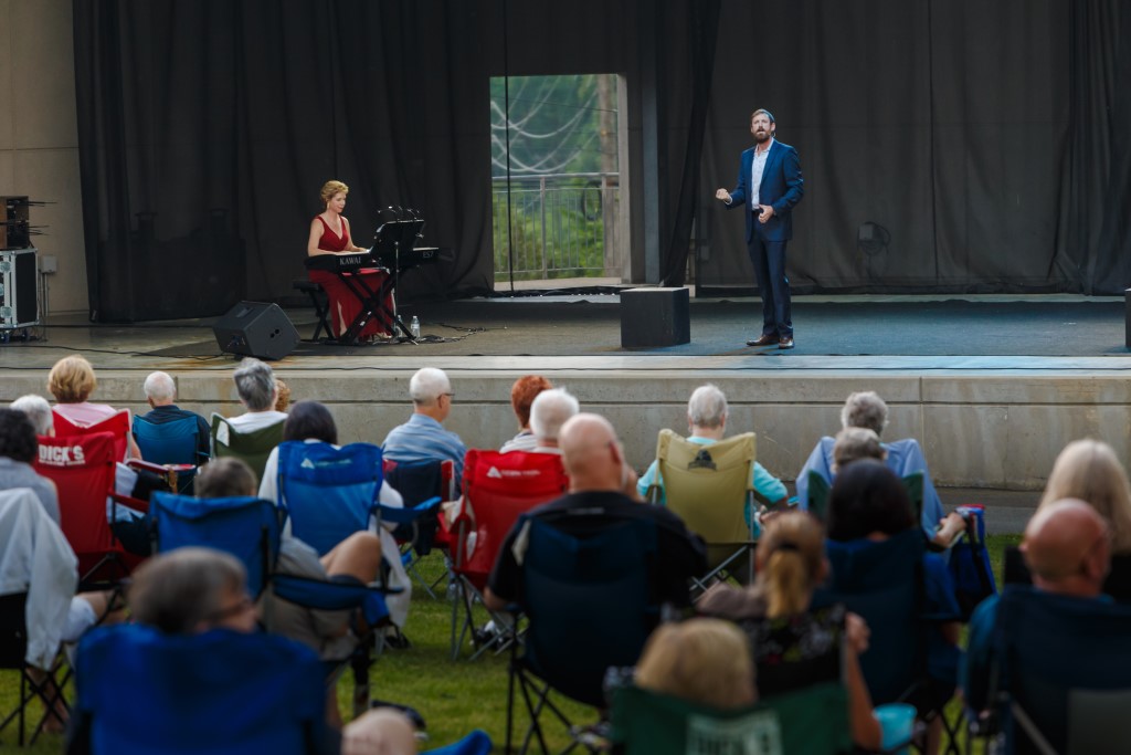 A baritone in a blue suit sings on the South Park Amphitheater stage as a pianist plays and lawn-chair audience listens