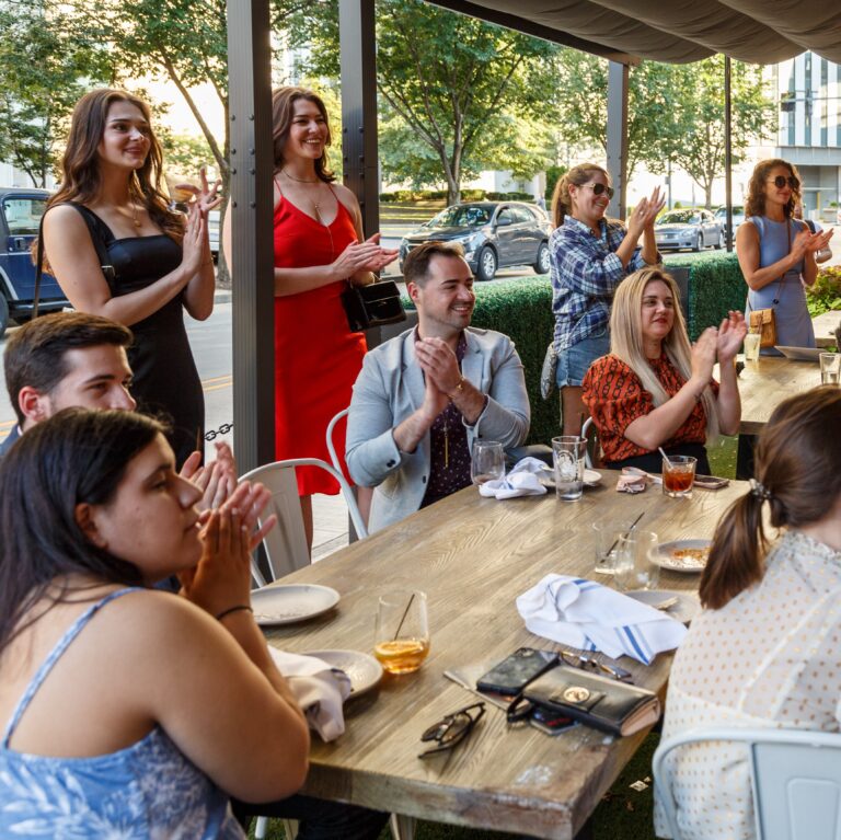 New Guard Summer Salon guests applaud around a long table on a covered patio