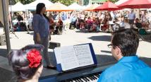 A pianist reads a score as a singer performs to a crowd under tents at the Market Square farmers market