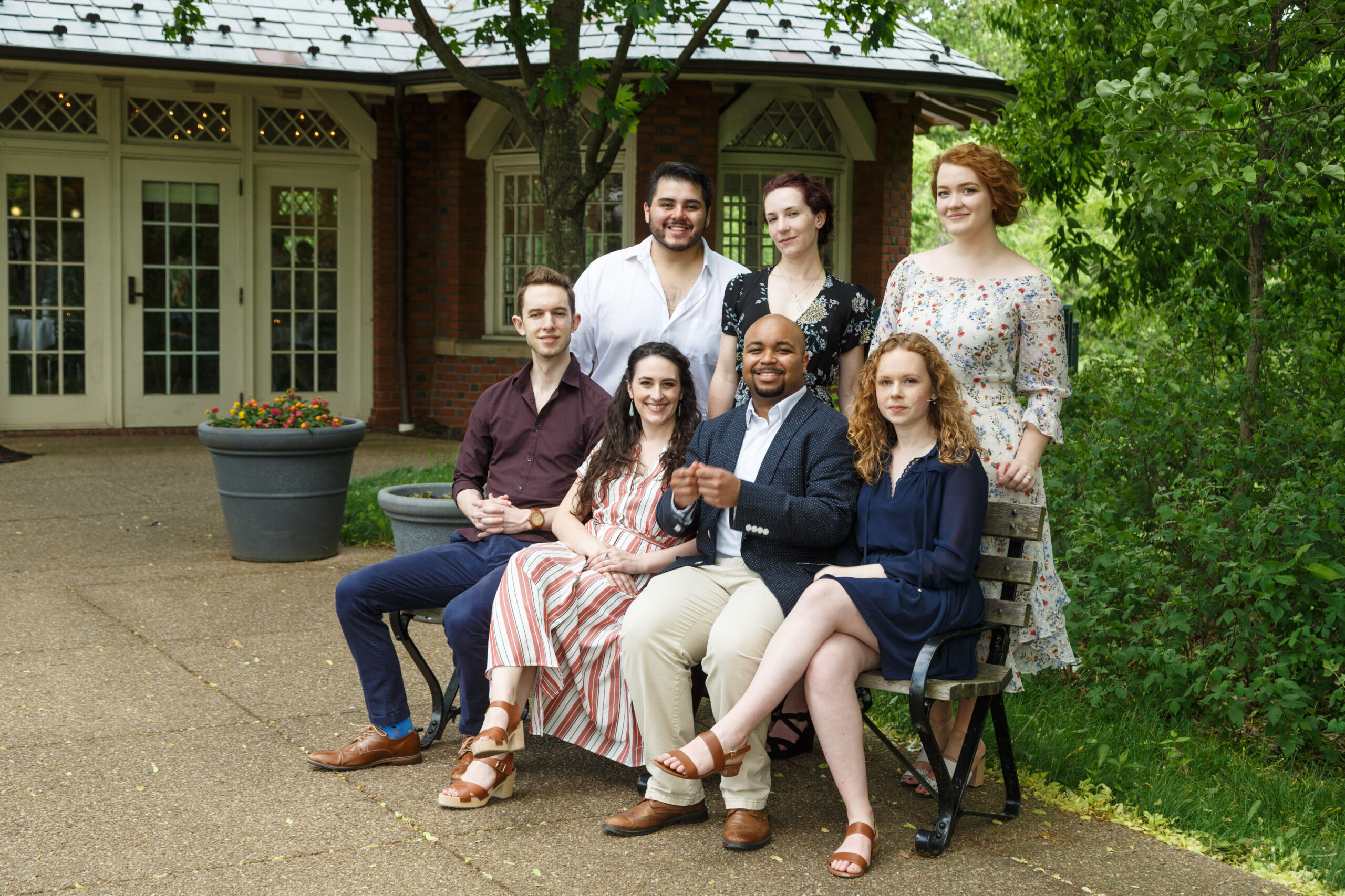 Seven Pittsburgh Opera Resident Artists pose on and around a park bench before a brick garden pavilion