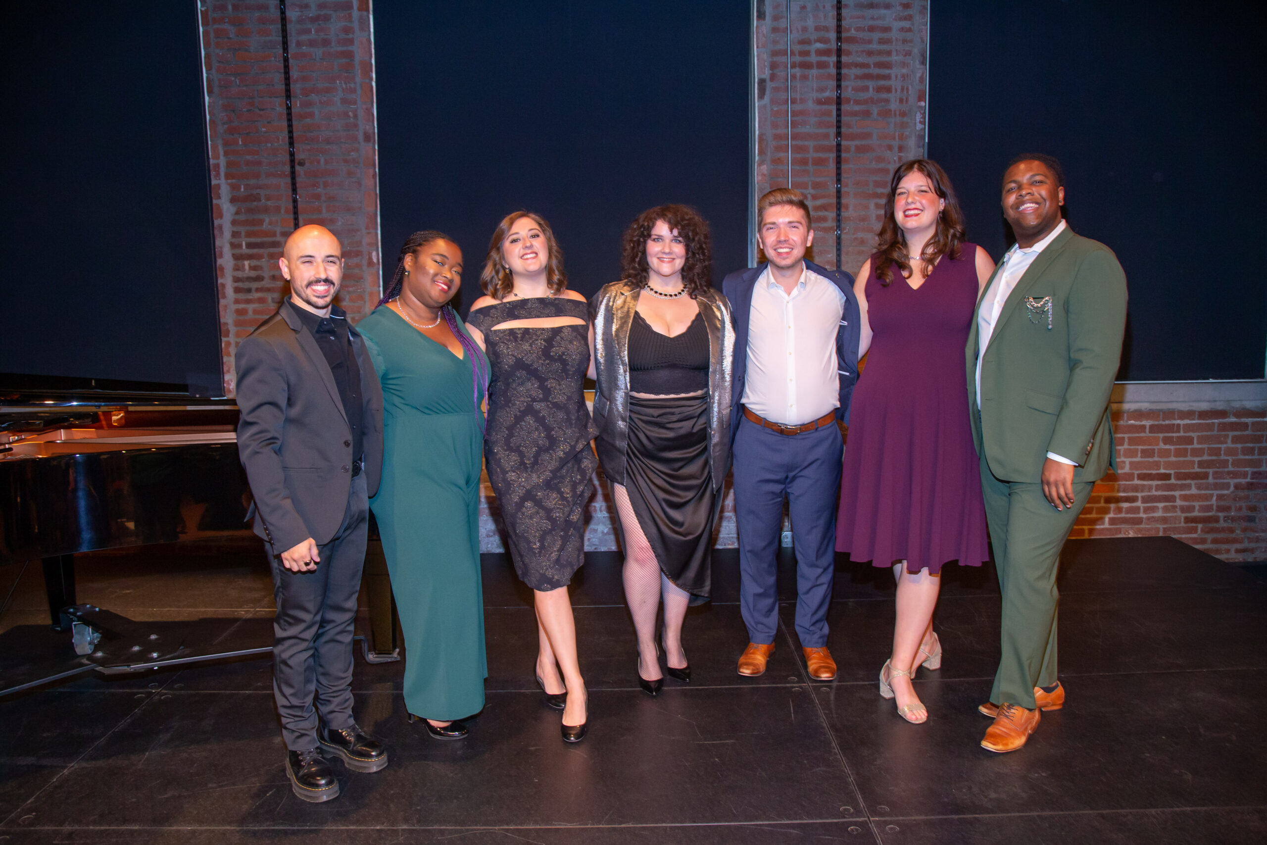 Seven performers in evening wear beside a grand piano on a brick-walled stage at a Pittsburgh Opera Rising Stars concert