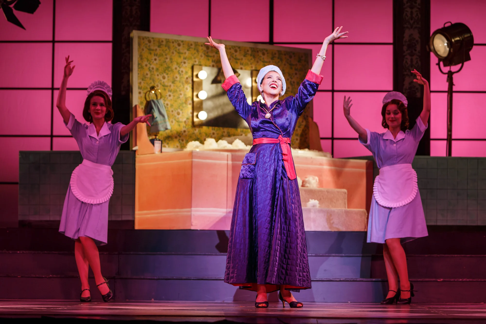 A woman in a purple satin robe raises both arms as two maids dance in a pink bathroom set in Pittsburgh Opera's Don Pasquale