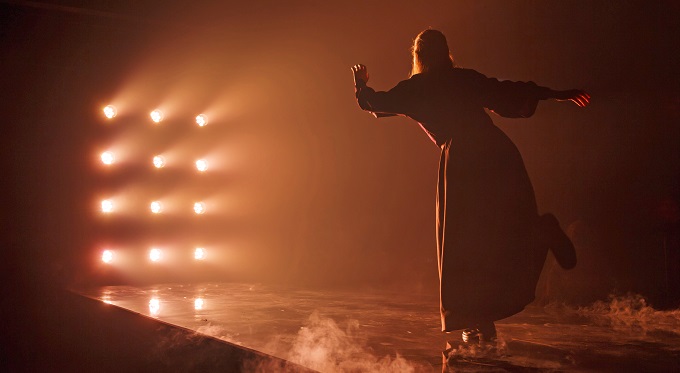 A robed figure in silhouette reaches toward a grid of glowing lights through haze in Pittsburgh Opera's In a Grove
