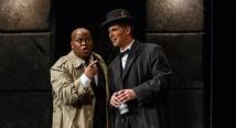 Leporello in a suit sings into a microphone beside Don Giovanni in a bowler hat in Pittsburgh Opera's Don Giovanni