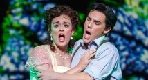 Rosalba in green and Arcadio in a blue vest sing together before a floral backdrop in Florencia en el Amazonas