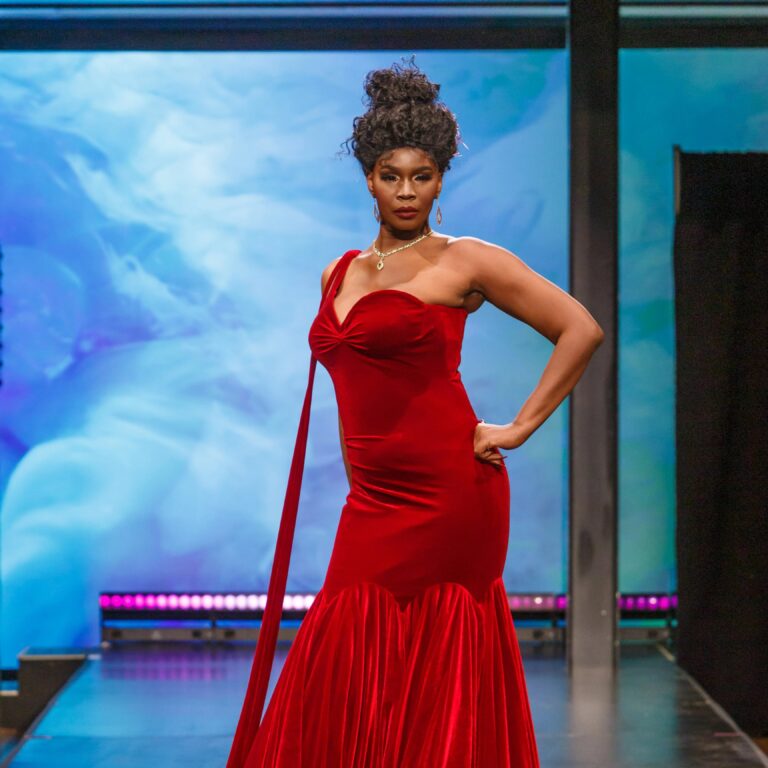 A model in a red velvet one-shoulder gown poses on the runway before a blue projection at the 2024 Fashion Show