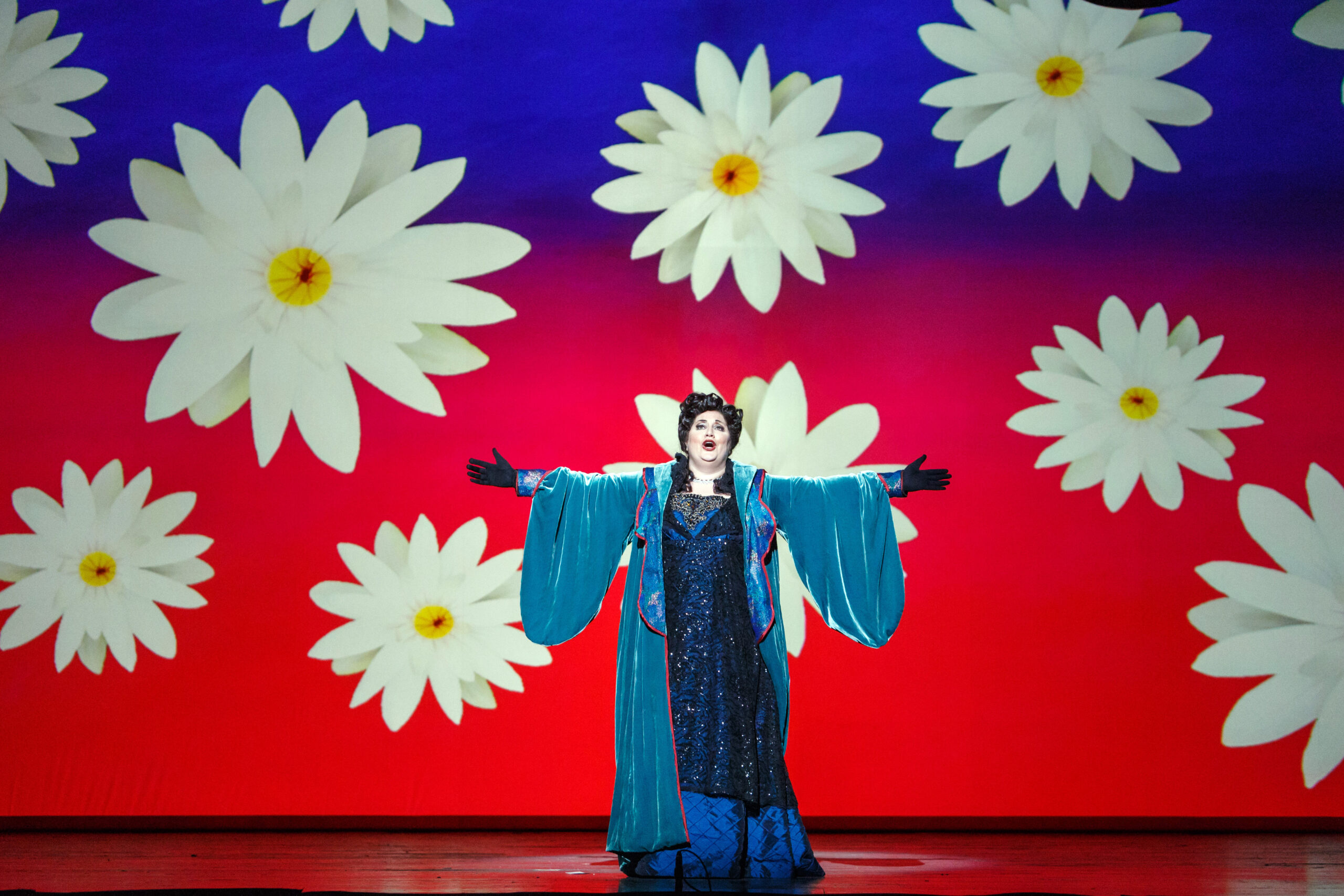 Florencia in a teal robe spreads her arms before a wall of giant daisies in Pittsburgh Opera's Florencia en el Amazonas
