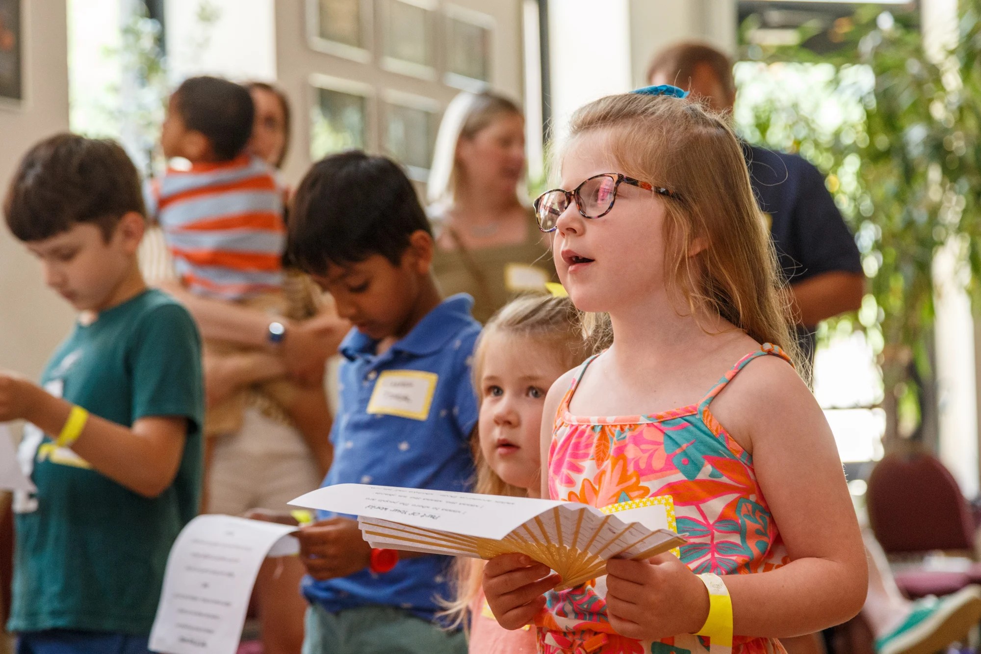A young girl in glasses and a floral dress sings from a paper fan as other children read lyrics at Family Day 2024