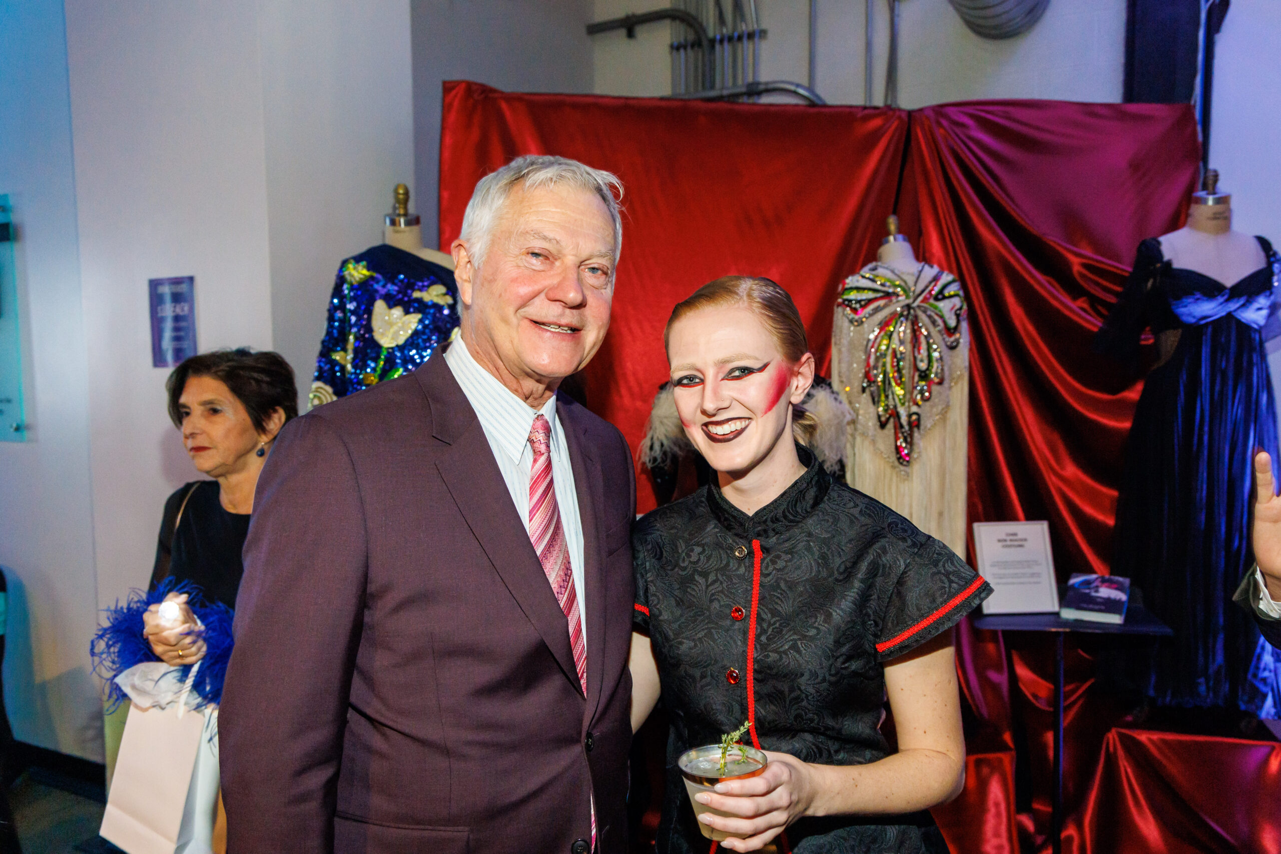 Christopher Hahn in a plum suit poses with a costumed guest before sequined gowns at the It's About Time fashion show