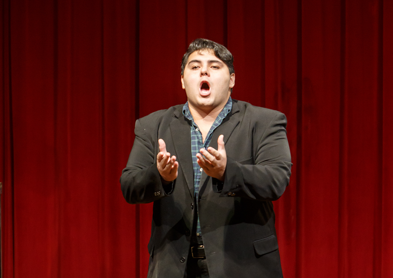 Angel Romero in a black blazer and plaid shirt sings with open hands before a red curtain at a Brown Bag concert