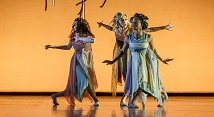 Three dancers in flowing green and gold costumes pose on an orange-lit stage in Pittsburgh Opera's Ariodante