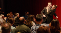 A tenor and a soprano embrace while singing before a seated audience at a Brown Bag concert