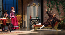 Rosina in a red gown stands as Bartolo reclines on a settee in Pittsburgh Opera's The Barber of Seville, 2023