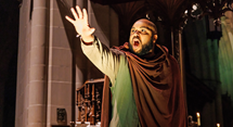 The Ferryman in a hooded robe raises a hand in a church in Pittsburgh Opera's Curlew River