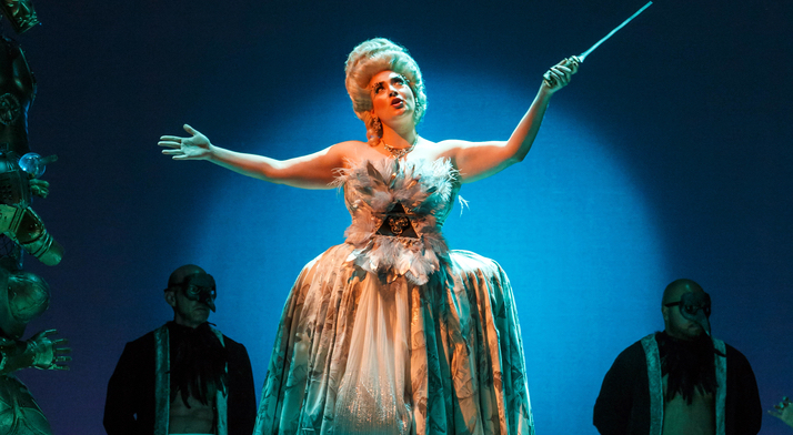 Alcina in a towering white wig and feathered gown raises a wand as masked servants flank her in Pittsburgh Opera's Alcina