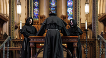 Three hooded monks gather at a wooden altar before stained glass in Pittsburgh Opera's Curlew River