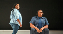 Two singers in denim, one seated, on a bare stage in Pittsburgh Opera's Denis & Katya