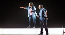Two singers in denim stand on a bare white stage in Pittsburgh Opera's Denis & Katya