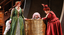Two women in green and red period gowns stand over a laundry basket with a man peeking out, in Pittsburgh Opera's Falstaff