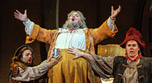 Falstaff in a gold doublet raises his arms between two followers in Pittsburgh Opera's Falstaff