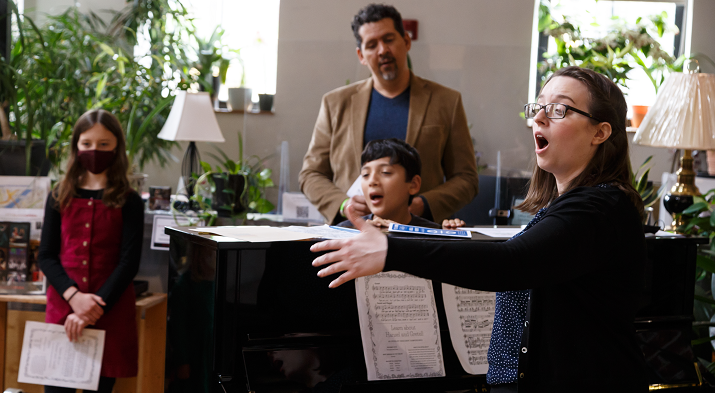 A soprano in glasses sings beside a grand piano as a boy and a masked girl sing along at Family Day 2022