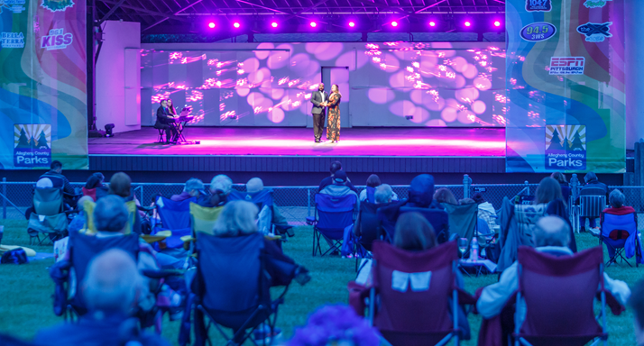 Two singers perform on the Hartwood Acres Amphitheater stage under purple light as a lawn-chair audience watches
