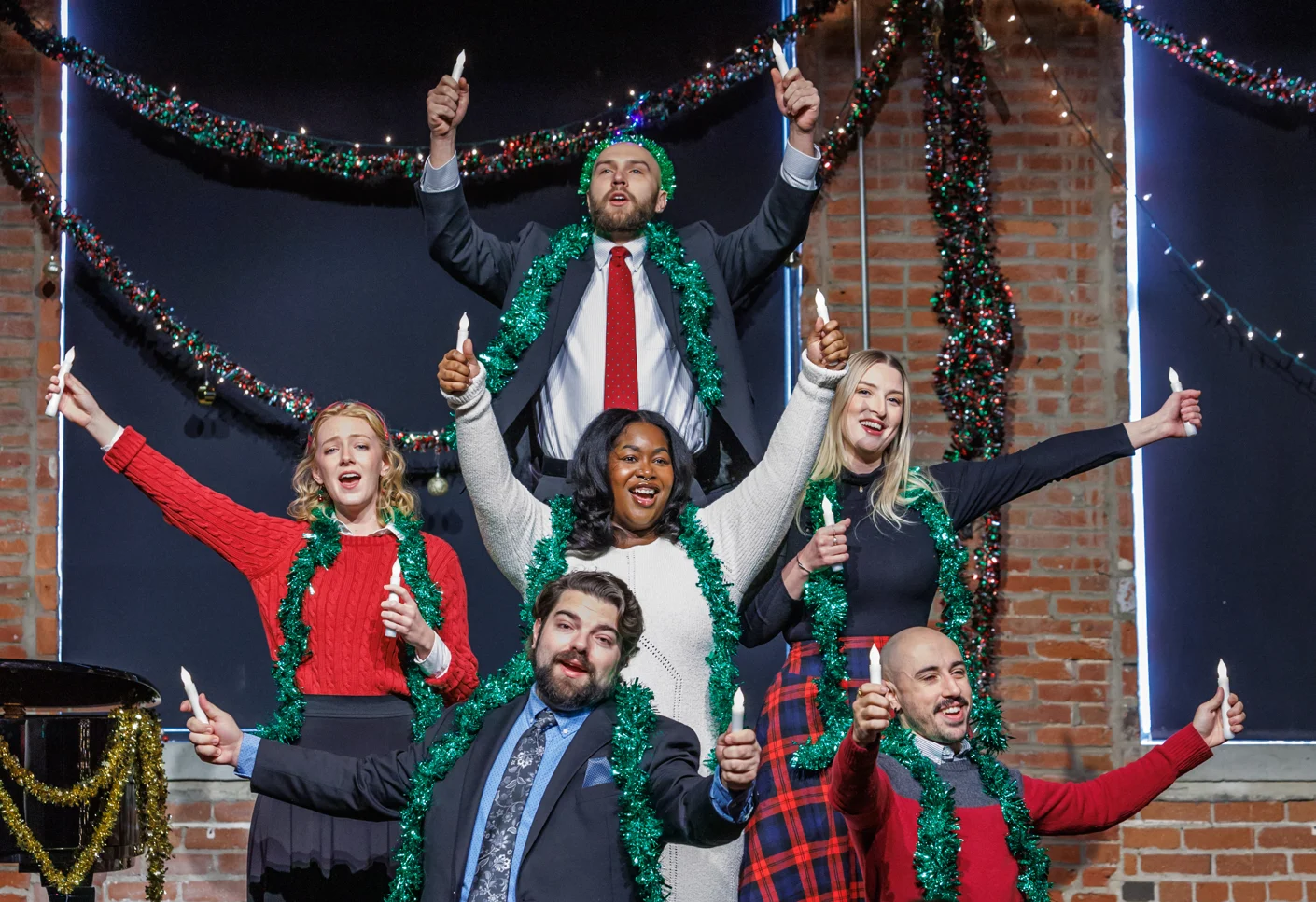Six Resident Artists in tinsel garlands hold candles aloft before holiday lights at the Holiday Song Shop