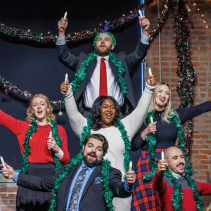 Six Resident Artists in tinsel garlands hold candles aloft before holiday lights at the Holiday Song Shop