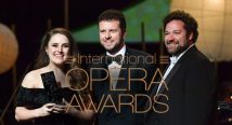 Three people hold an award on stage before the International Opera Awards logo