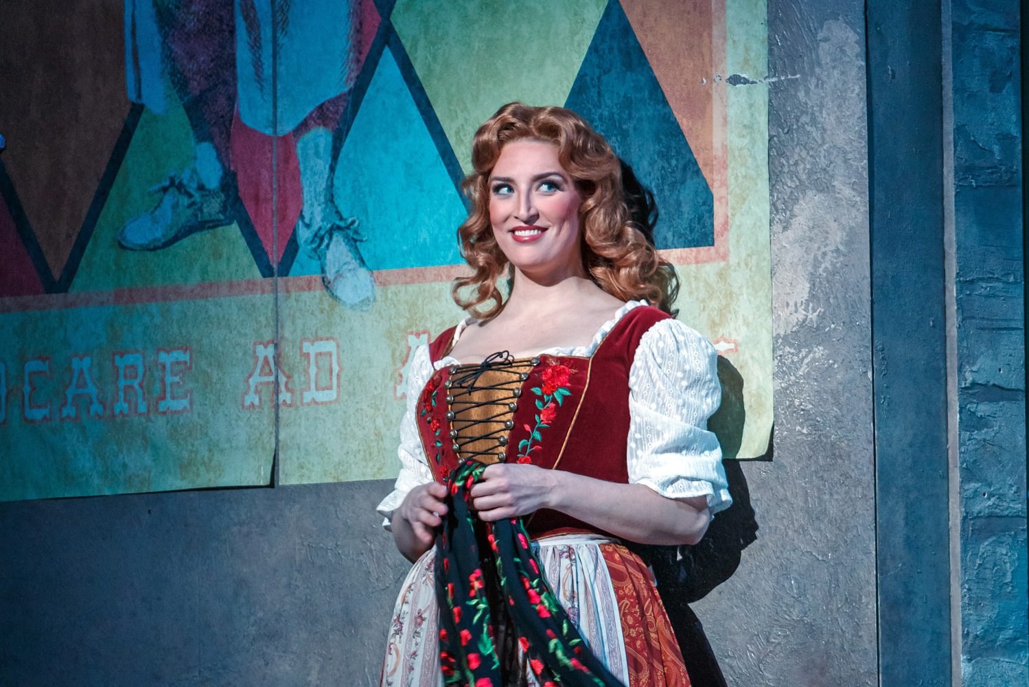 A singer in a red laced bodice and auburn curls smiles beside a painted poster on stage