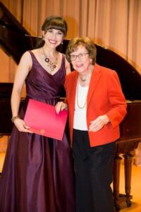 Shannon Jennings in a plum gown holds a red certificate beside Mildred Miller Posvar in a red jacket by a piano