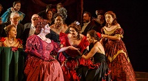 Violetta in a red gown raises a glass as party guests dance behind her in Pittsburgh Opera's La Traviata, 2024
