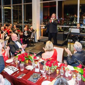 A tenor in a red bow tie sings to applauding guests at red-draped tables at Maecenas, city lights behind