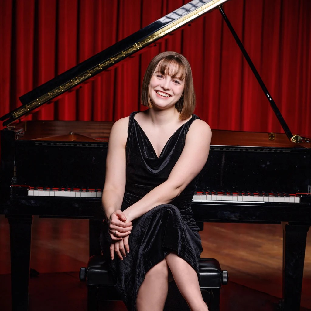 Maeve Berry seated at a grand piano before a red curtain, portrait