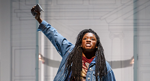 A singer in a denim jacket raises a fist holding a phone in Pittsburgh Opera's We Shall Not Be Moved