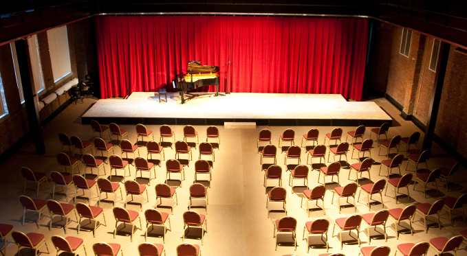 Rows of empty chairs face a grand piano on a stage before a red curtain in the George R. White Opera Studio