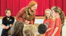 A red-haired presenter in a patterned shawl talks with children in red T-shirts in a school hall
